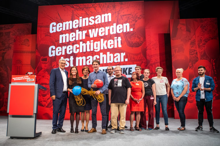 LINKE Mitglieder stehen vor einer Parteitagswand auf der in großen Lettern "Gemeinsam mehr werden" steht.